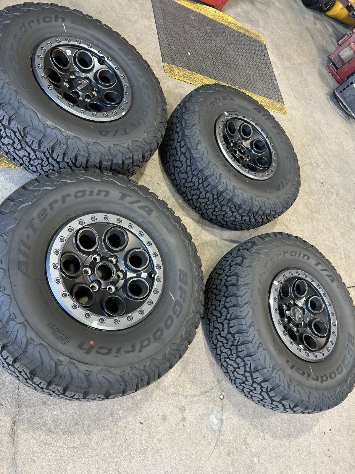 Four off-road wheels in matte black with gunmetal beadlock rings, mounted on all-terrain tires